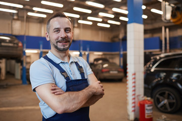 Waist up portrait of smiling car mechanic standing with arms crossed while posing in auto repair...