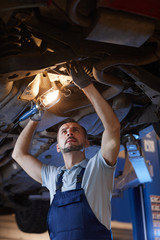 Low angle portrait of mature mechanic looking under car on lift and holding lamp light during inspection in auto repair workshop, copy space