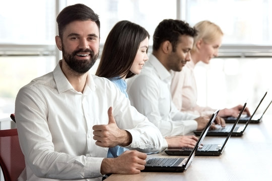 Smiling cheerful caucasian bearded man with thumb up in office. Young adult man with beard smiling with manager, accompanied with colleagues and shows thumb up.