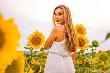 Lifestyle, blonde girl among sunflowers in a white dress and a belt looking at camera