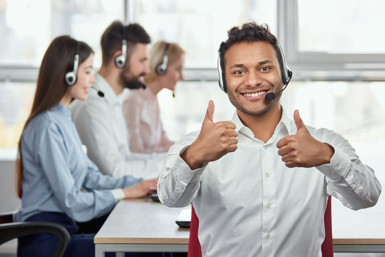 Indian man shows two thumbs up against bright office background. Happy call centre worker giving a thumbs up and looking to camera in front of working people in office backgorund. - Powered by Adobe