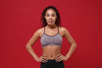 Smiling young african american sports fitness woman in sportswear posing working out isolated on red background in studio. Sport exercises healthy lifestyle concept. Stand with arms akimbo on waist.