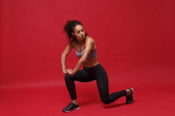 Fototapeta na wymiar Attractive young african american sports fitness woman in sportswear posing working out isolated on red wall background. Sport exercises healthy lifestyle concept. Stretching her legs, doing lunges.