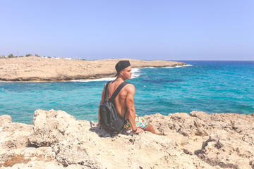 Fototapeta na wymiar Athletic young man in cap sitting around Natural Rock Bridge at Cape Greco near Ayia Napa in the evening light. Cyprus. Healthy lifestyle