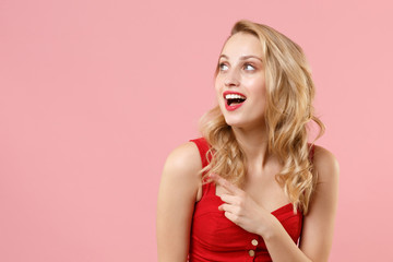 Excited young blonde woman girl in red sexy clothes posing isolated on pastel pink wall background studio portrait. People emotions lifestyle concept. Mock up copy space. Pointing index finger aside.