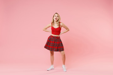 Funny young blonde woman girl in red sexy clothes posing isolated on pastel pink background studio portrait. People emotions lifestyle concept. Mock up copy space. Standing with arms akimbo on waist.