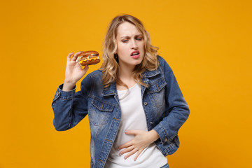 Sick young woman in denim clothes isolated on yellow orange wall background studio portrait. Proper nutrition or American classic fast food concept. Hold burger put hand on stomach, having ache pain.