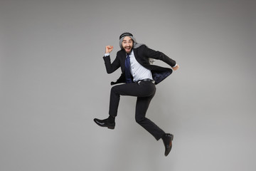 Fototapeta na wymiar Cheerful young bearded arabian muslim businessman in keffiyeh kafiya ring igal agal classic black suit isolated on gray background. Achievement career wealth business concept. Jumping like running.