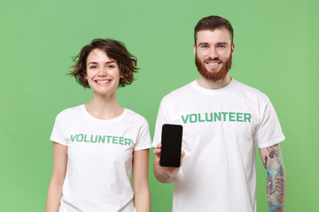 Smiling two young friends couple in white volunteer t-shirt isolated on green background. Voluntary free work assistance help charity grace teamwork concept. Hold mobile phone with blank empty screen.