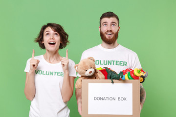 Excited two friends couple in volunteer t-shirt isolated on green background. Voluntary free work assistance help charity grace teamwork. Hold donation box with kids toys pointing index fingers aside.