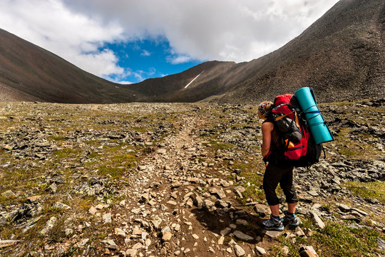 A tourist with a large backpack stands and looks at a mountain pass. Rocky trail. Clouds over the mountains. Horizontal.