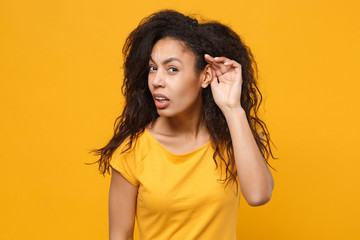 Dissatisfied curious young african american woman girl in casual t-shirt posing isolated on yellow orange background. People lifestyle concept. Mock up copy space. Try to hear you with hand near ear.