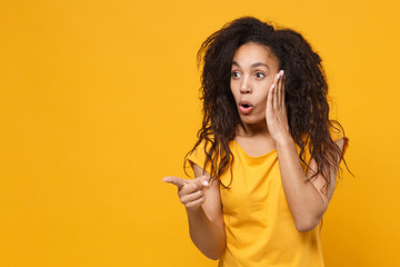 Shocked young african american woman girl in casual t-shirt posing isolated on yellow orange background. People lifestyle concept. Mock up copy space. Pointing index finger aside, put hand on cheek.