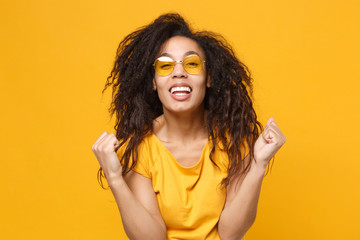 Joyful young african american woman girl in casual t-shirt, eyeglasses posing isolated on yellow orange background studio portrait. People lifestyle concept. Mock up copy space. Doing winner gesture.