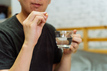 Young Asian man feeling sick and taking pills while sitting on the bed at home with medicines tablets and drinking water on table. Healthcare and Medicine concept