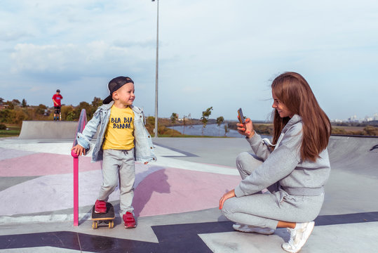 Woman mom photographs phone, little boy son 3-5 years old, learning to ride skateboard, summer on sports ground, emotions happiness, fun, relaxation pleasure. Record video on a smartphone.