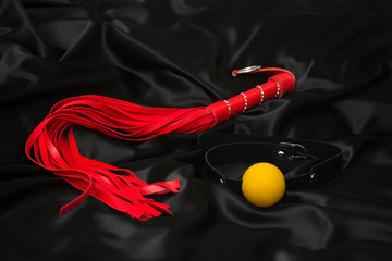 Red whip and gag on a black silk background. Accessories for adult sexual games