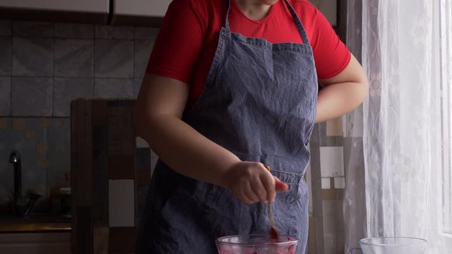 beautiful mature woman in red sweater and gray economic apron prepares salad of boiled beets, garlic and mayonnaise. girl rubs the beets, squeezes garlic and season with mayonnaise