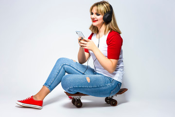 Happy Caucasian Female Blond Girl In Casual Closing Listeining To Music in Headphones On Smartphone. posing on Skateboard Against White.