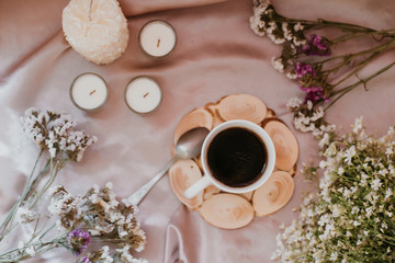 Cup of coffee on a stand surrounded by flowers and candles of different sizes and shapes