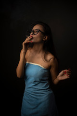 Portrait of Asian/Japanese/Korean/Indian brunette young girl in blue bath towel  and spectacles smoking cigarette in black studio copy space background. Indian lifestyle and fashion photography