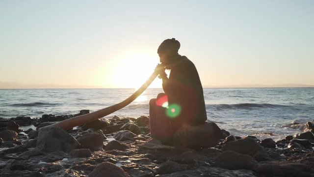 Silhouette of man playing didjireedoo in sunshine at seashore