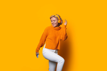 Caucasian blonde girl using headphones and wearing a nice sweater is dancing on a yellow background