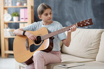 Content teenage girl sitting on sofa and using internet on tablet while learning to play guitar