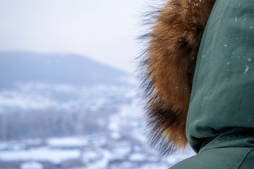 Man in a green hood in the winter. View from the back, fur. Valley in the mountains in blur. White snow and snowflakes.