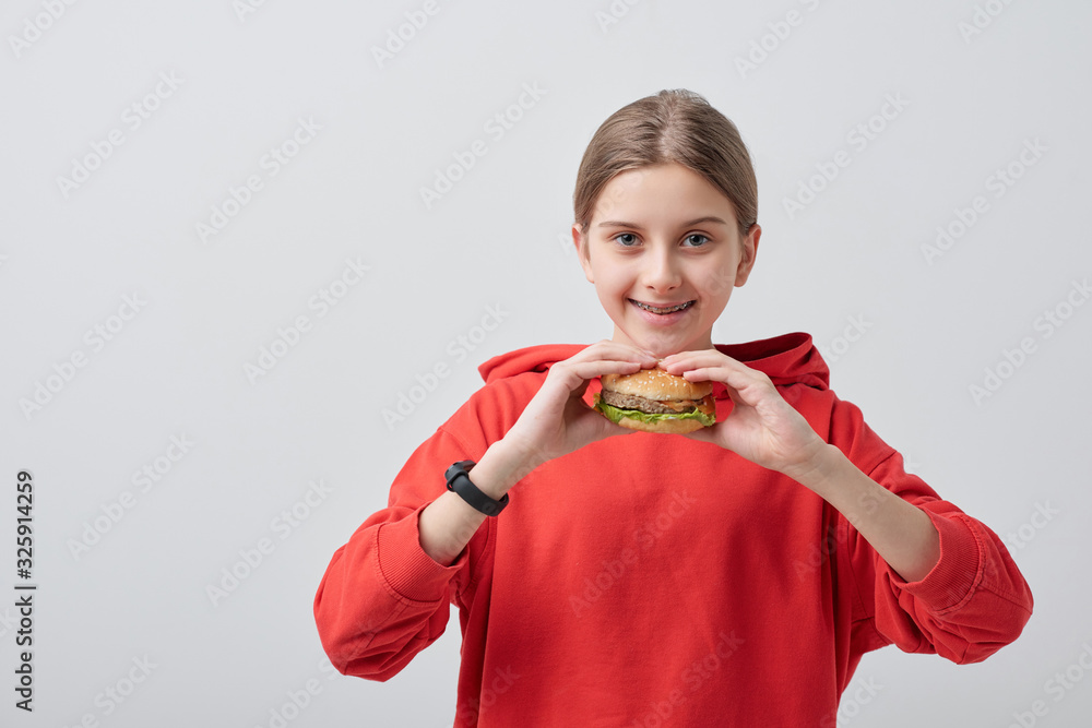 Wall mural portrait of smiling teenager in red sweatshirt eating burger against white background