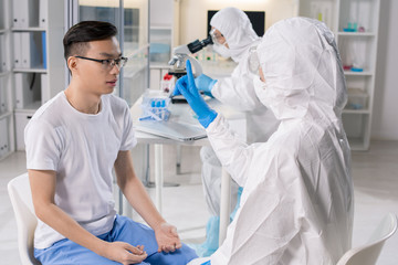 Medical specialist in protective suit showing finger to infected Asian patient while checking his reaction