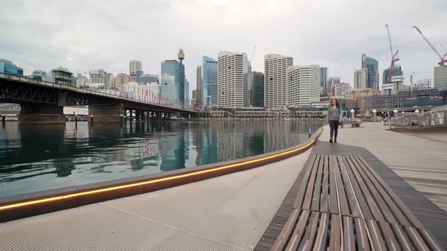 A female tourist walks up to a bench before sitting down and admiring the Sydney CBD from Darling Harbour