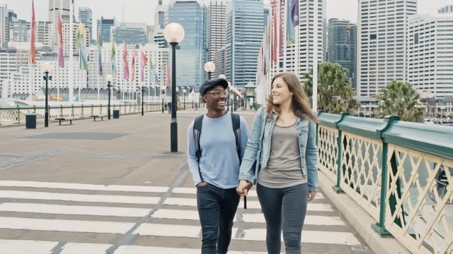 Smiling multiracial couple walk across Pyrmont Bridge, Darling Harbour, as the Sydney CBD encompasses the background.
