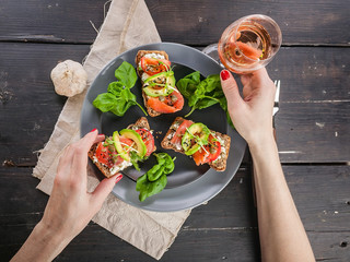 Seafood and Rose Wine. Woman eating tasty bruschettas with smoked salmon, curd cheese, avocado, basil and chia seeds. Tasty and healthy snack. Mediterranean Kitchen. Close up. Top view
