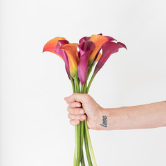 Close up cropped view of hand with love tattoo on wrist holding red and orange calla lilies...