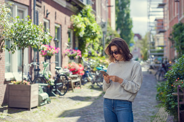 A young woman uses a smart phone while standing on a city street in the summer season. The concept of a lone tourist using a Navigator in a smart phone.