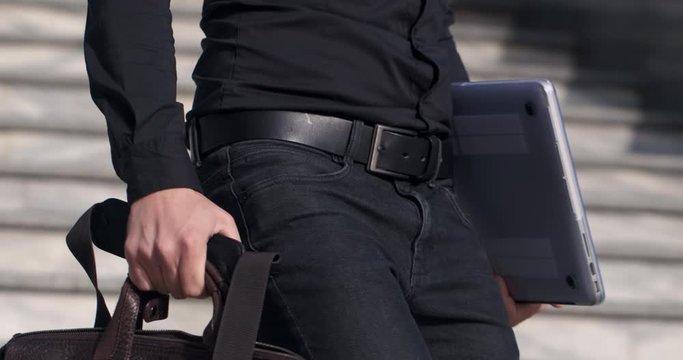 IT businessman in black clothes descends the stairs holding a bag in one hand and a laptop in the other.