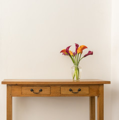 Red and orange calla lilies in glass vase on oak side table against white wall