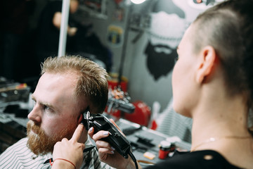  In a barber shop, a young woman cuts a guy with red hair and a red beard