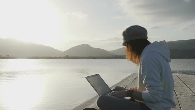 Internet freelance job choice concept: a young woman works on her laptop sitting on a pier by a lake at sunset