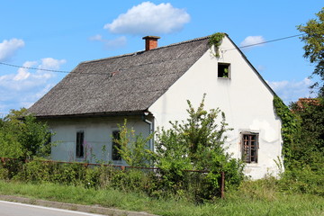 Fototapeta na wymiar Abandoned small white suburban family house with broken windows and dilapidated facade covered with old roof tiles surrounded with overgrown tall uncut grass and various vegetation next to paved road 