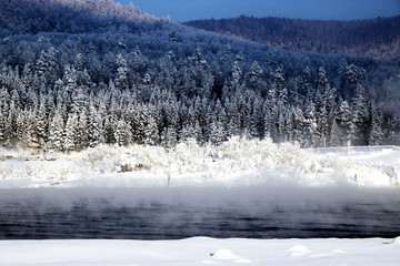 Fototapeta premium In the background, on the other bank of the river, conifer winter forest begins. Everything is simple, beautiful and expressive. In the foreground - mist rising from the water and fluffy snow.