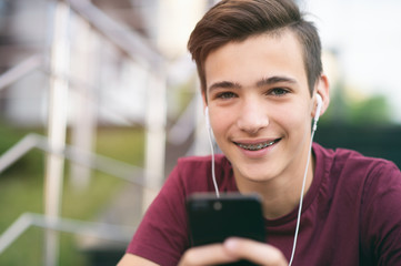 Close-up portrait of a smiling young man with a smartphone, in the street.  Happy teenage boy is using mobile phone, outdoors. Cheerful teenager spends time in social networks using cell phone. - obrazy, fototapety, plakaty