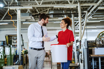 Handsome caucasian owner of printing shop dressed in shirt and tie talking to his new female employee. Woman holding bucket with liquid glue. Printing shop interior.