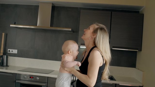 Young mother entertaining her son in the kitchen. Blond pregnant woman in black t-shirt tossing her little kid, smiling, laughing and higging him, standing near the desk with equipment. Male child