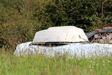 Plastic white small river boat taken from river and left flipped in family house backyard over winter next to stacked firewood surrounded with tall uncut grass and dense trees in background