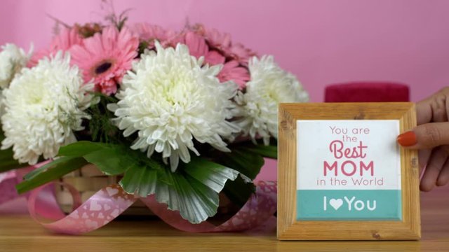 Closeup shot of a basket of pink and white flowers with a mother's day present. Woman hands placing a wooden frame with 'Best Mom' text next to a bouquet of flowers and a gift box - pink background