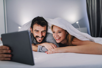 Young handsome cheerful caucasian couple lying on stomach on bed covered with bedsheets looking at tablet.