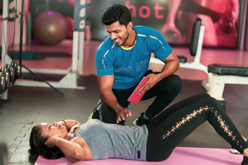 Trainer helping young woman to do abdominal exercises on the background of fitness balls. Beautiful woman doing abs on a mat and a man helps her in gym