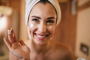 Young happy woman applying moisturizer on her face in the bathroom.